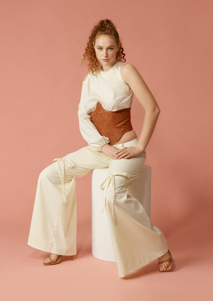 Model sitting on white modern circle stool wearing off white asyymetric blouse, off white wide leg cotton trouseres, and bourbon brown western saddle under bust corset. 