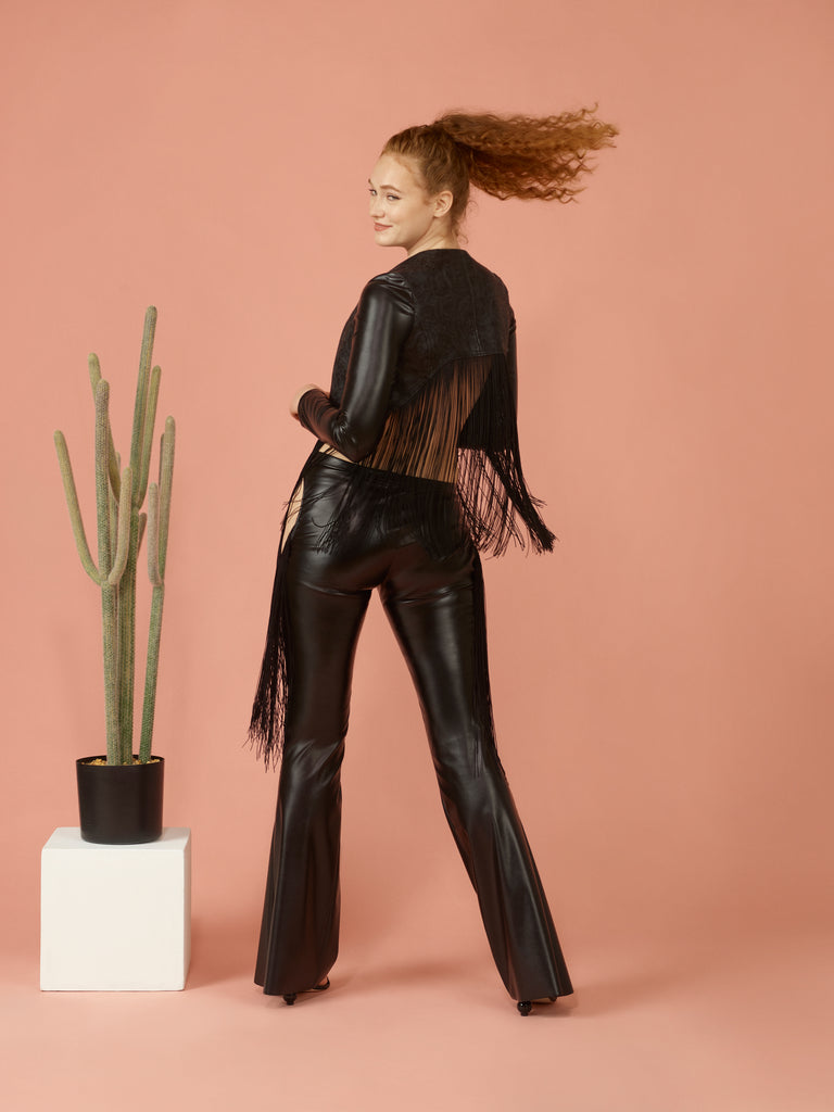 Back view of black western fringe jacket. Sleeves are a shiny black pleather. Back jacket hem is a high-low styled hem. Model is wearing black stretchy pleather jeans.